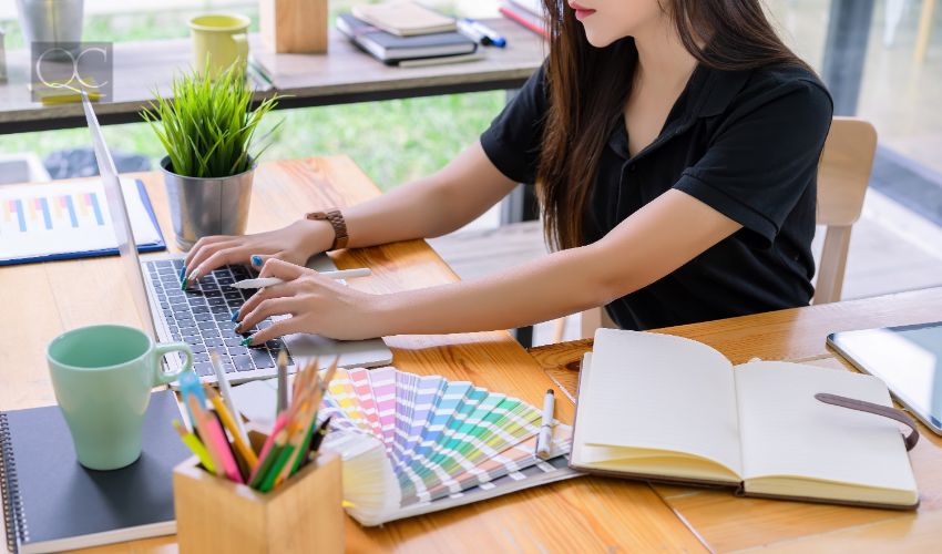 Color consultant certification in-post image, adult woman working on laptop at desk, with color swatches and notebook beside her