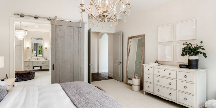 Beautiful master bedroom in new luxury home with view of ensuite master bathroom. Features barn door, chandelier, and elegant furnishings. Interior decorator article.