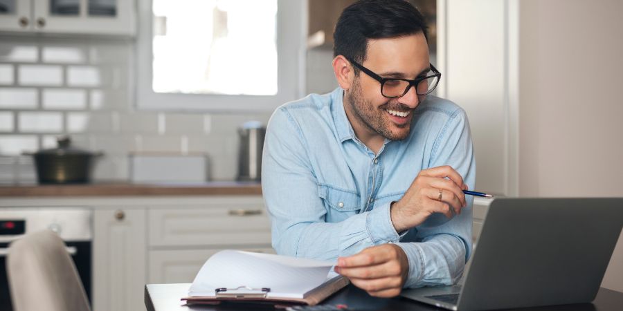 Young man working from home doing paperwork while using laptop and holding pen in his hand. Stay motivated online course article.